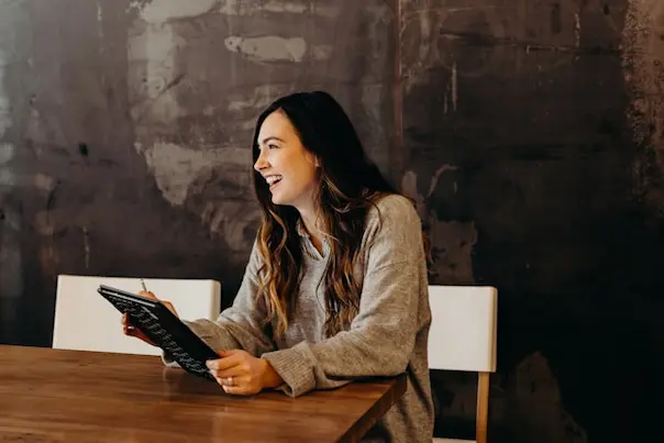 Happy woman smiling sitting down on a chair holding a book and pen over a table.